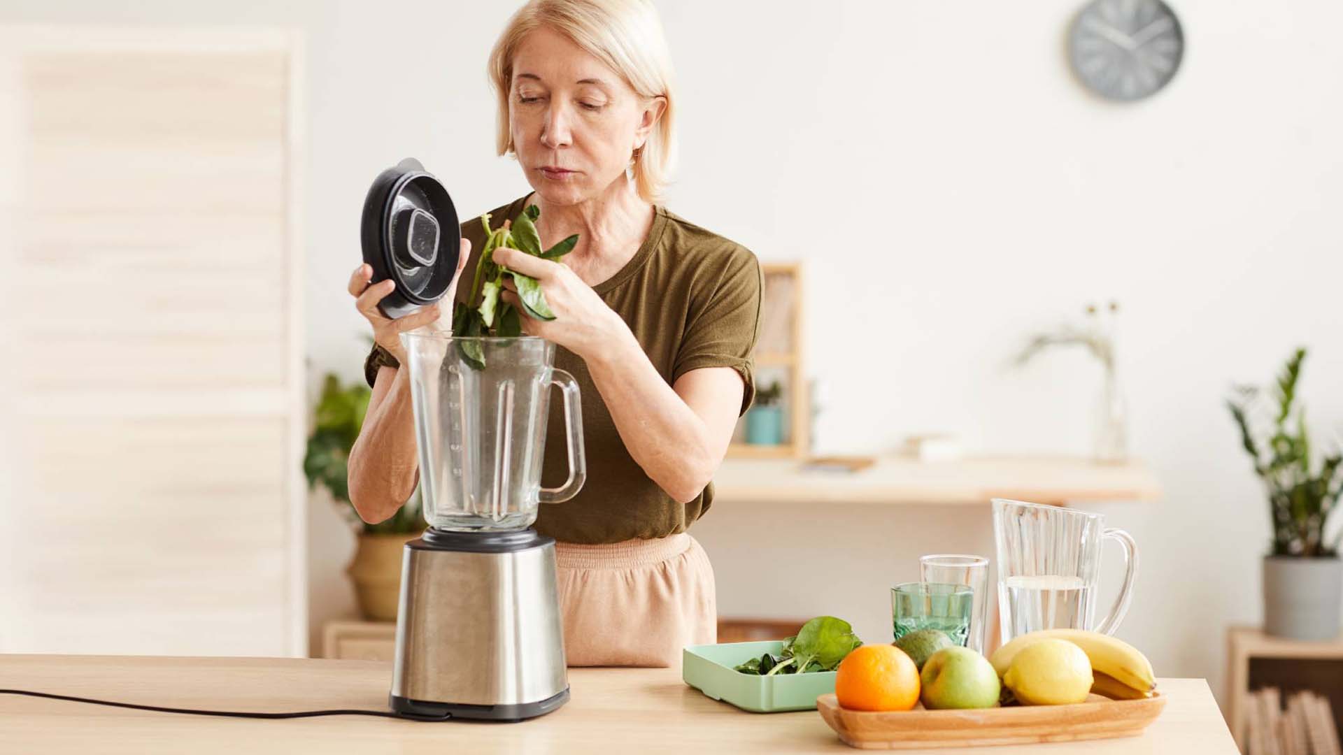 Mature woman making healthy cocktail from spinach and fruits using blender while standing in the kitchen