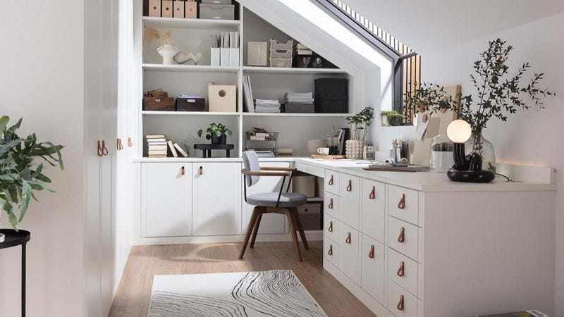 A modern home office with large built-in desk, drawers and wall shelving in white, with black furnishings such as a desk chair and side table