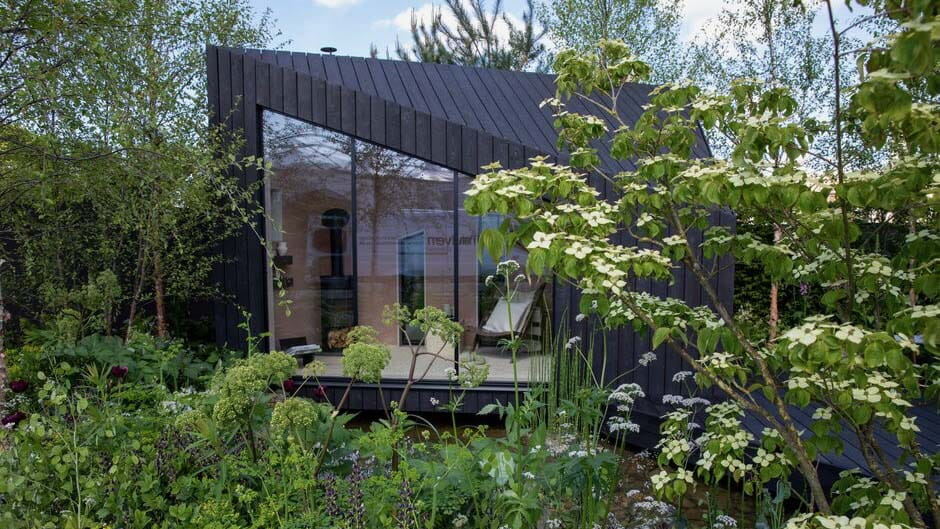 A small carbon-neutral garden cabin made of dark grey strips with a large glass front is situated at the center of a wild, green garden