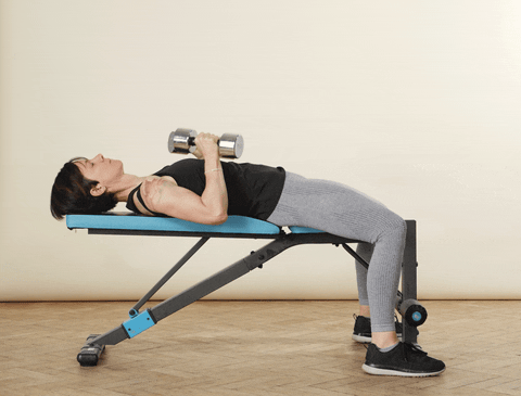 A woman in sports clothing lying on an exercise bench with feet on the ground either side, demonstrating a dumbbell chest press