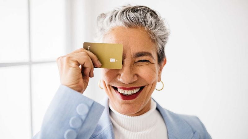 Mature woman in a blue blazer, standing near a window holds a gold credit card over her left eye