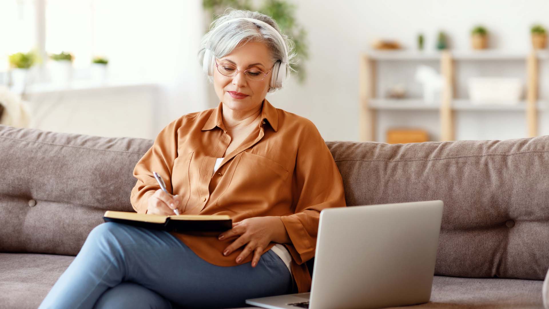 Silver haired older lady writing on a note pad with headphones on