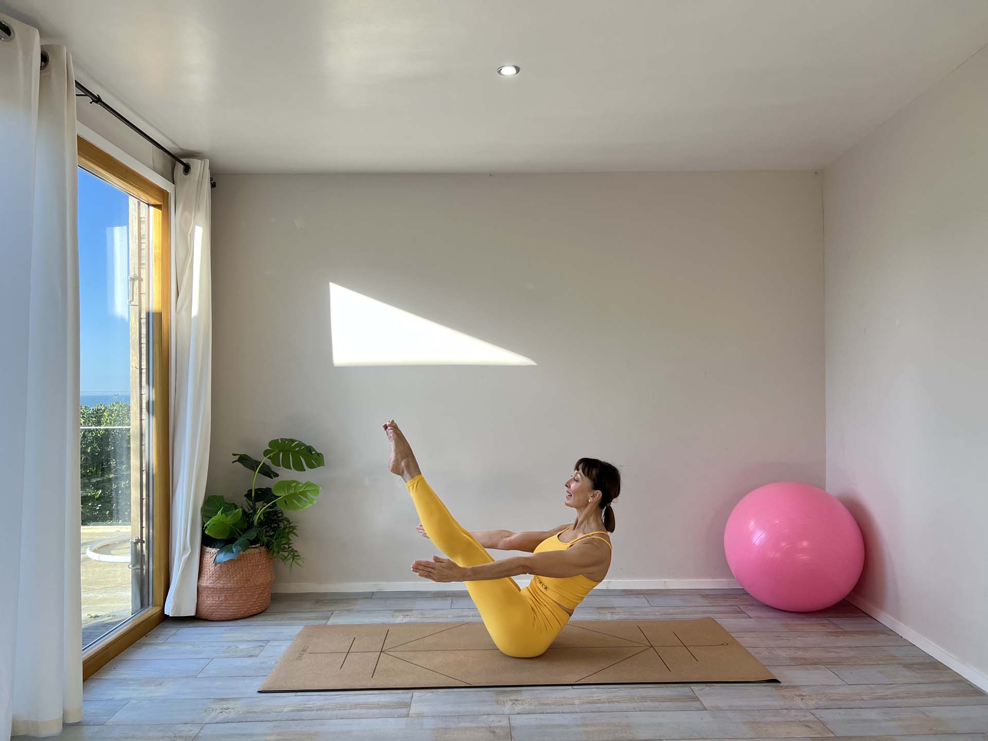 A woman holding a Pilates pose in a studio with the sun streaming in through patio doors.