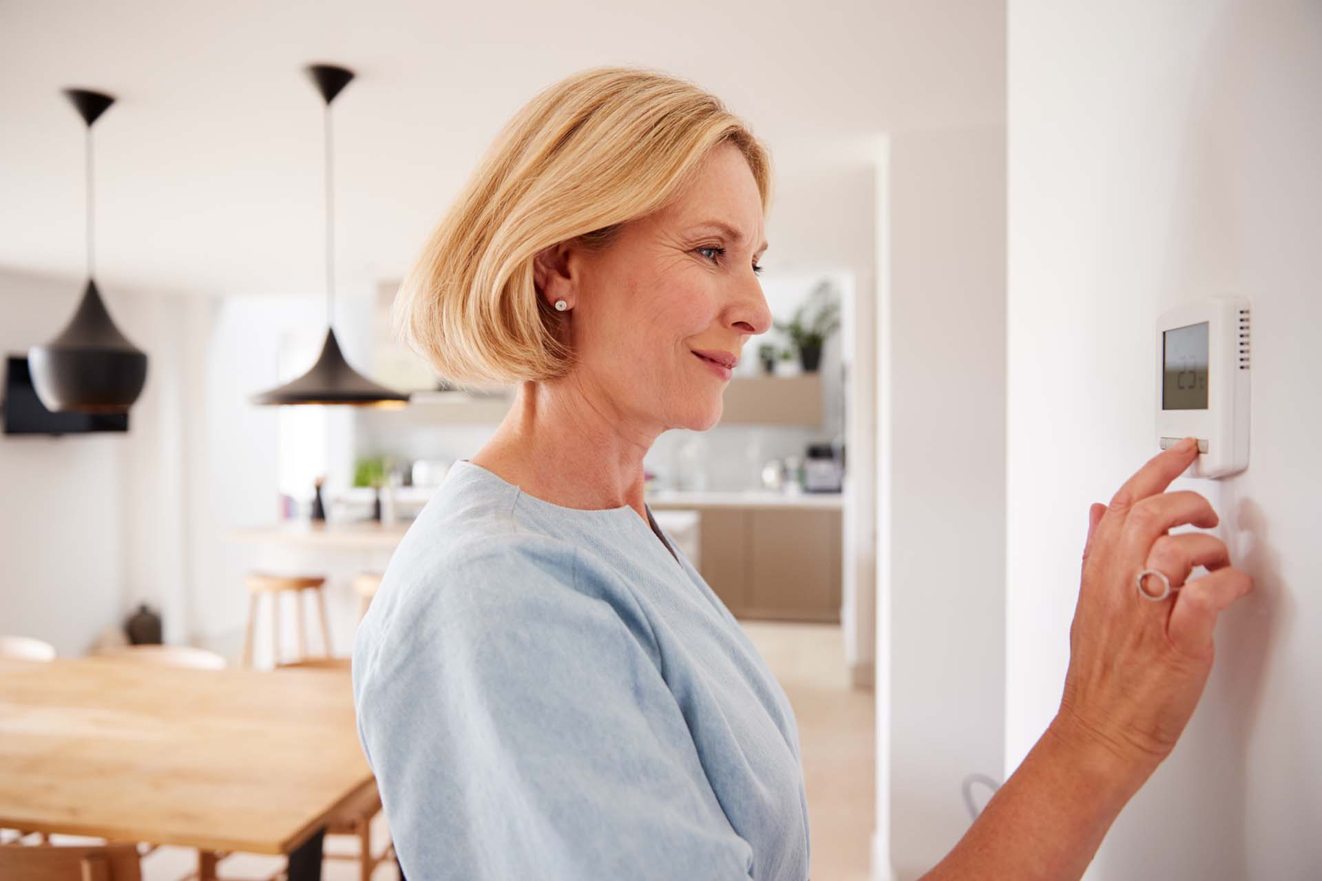 A woman in a blue top with a blonde bob adjusting a thermostat with a kitchen setting visible in the background