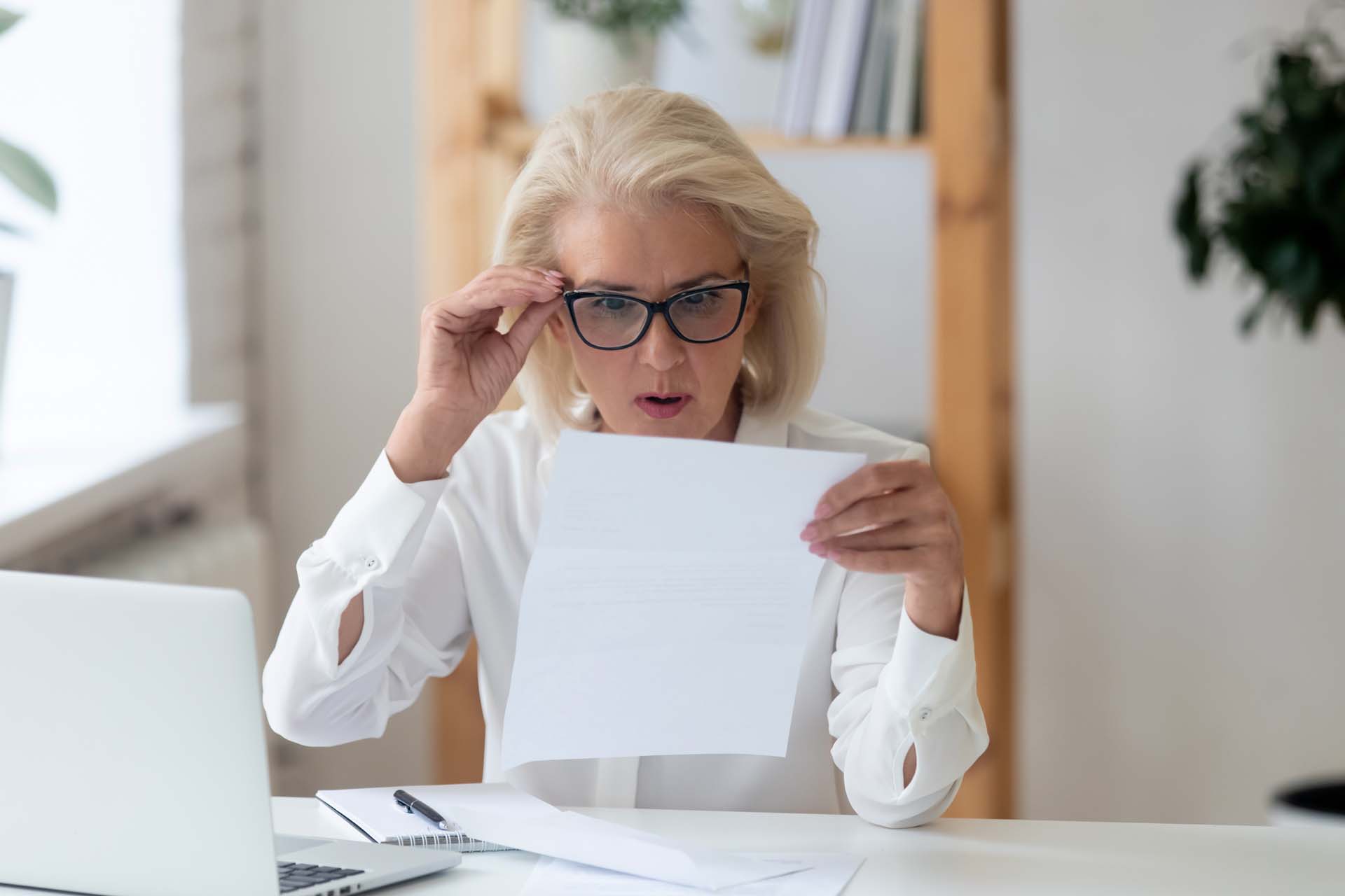 A blonde woman wearing glasses looks shocked while reading from a piece of paper