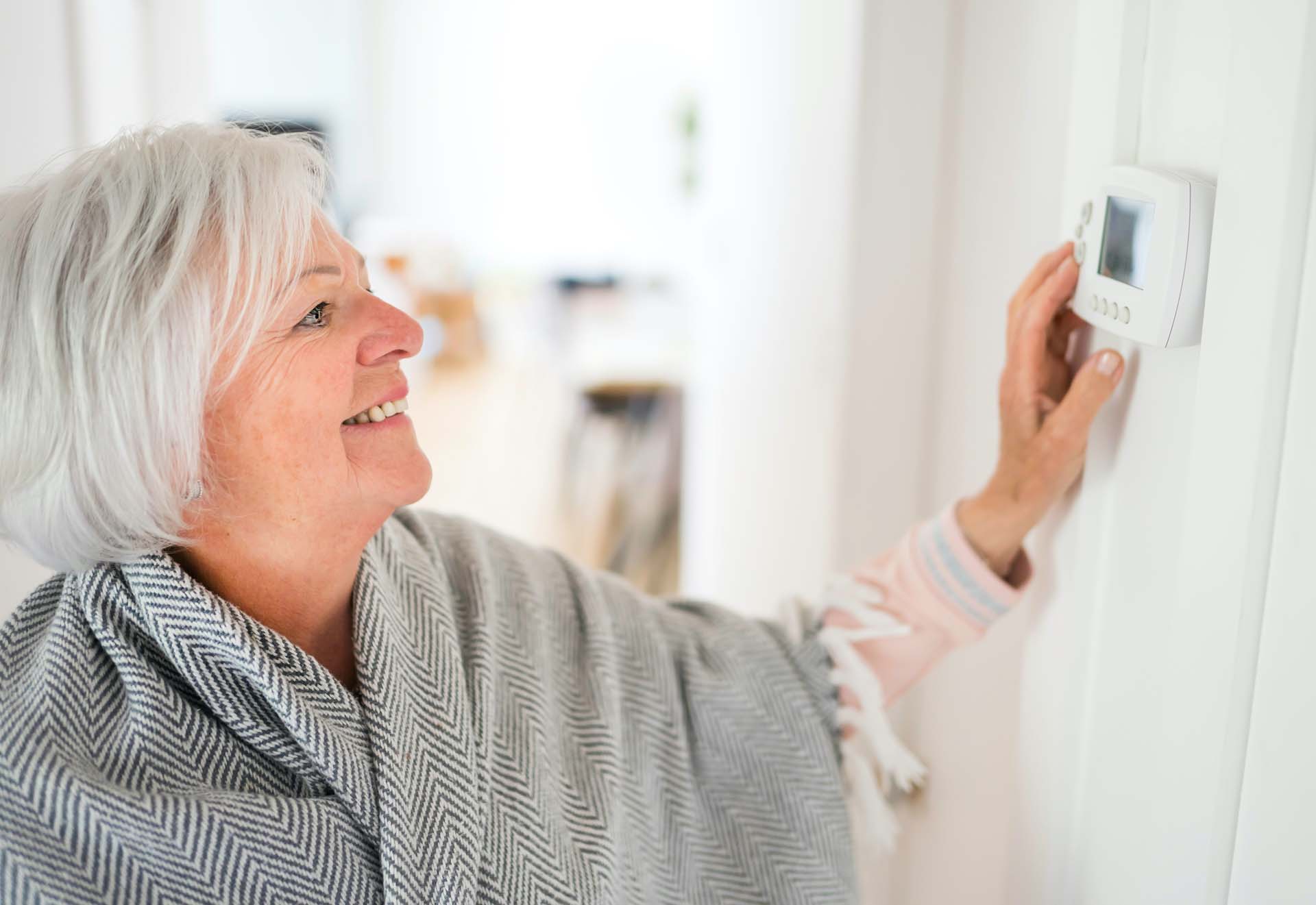 A mature woman wearing a grey shawl adjusts a wall thermostat