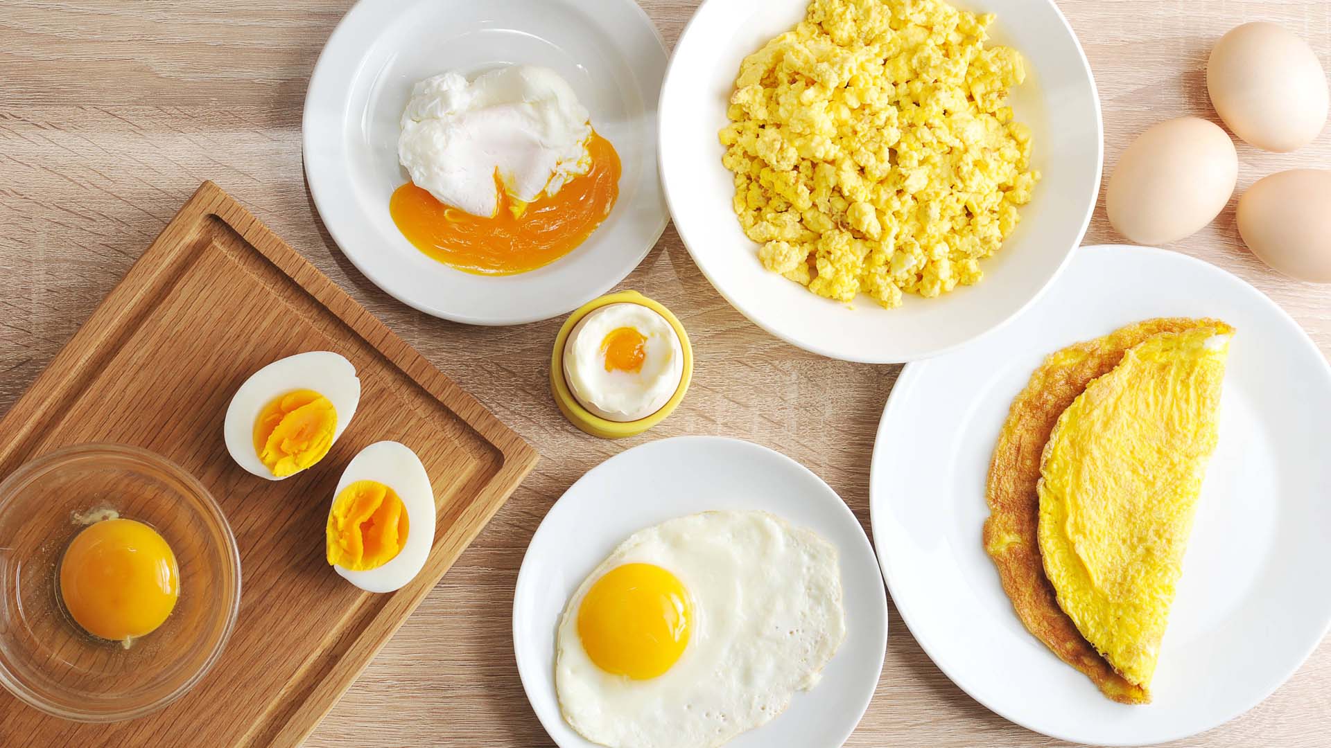 A flat lay image of eggs cooked in different ways, including a bowl of scrambled eggs, an omelette, fried egg, poached egg with the yolk spilling, a hard boiled egg in halves and a raw egg yolk
