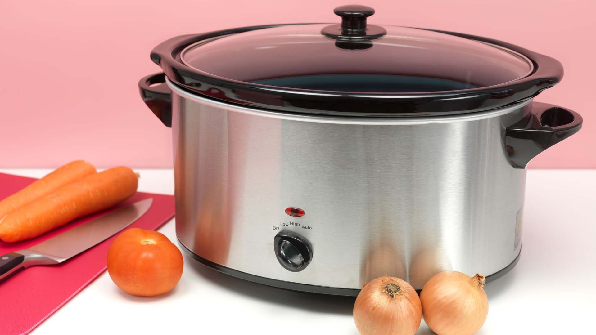 An electric slow cooker with vegetables and a chopping board