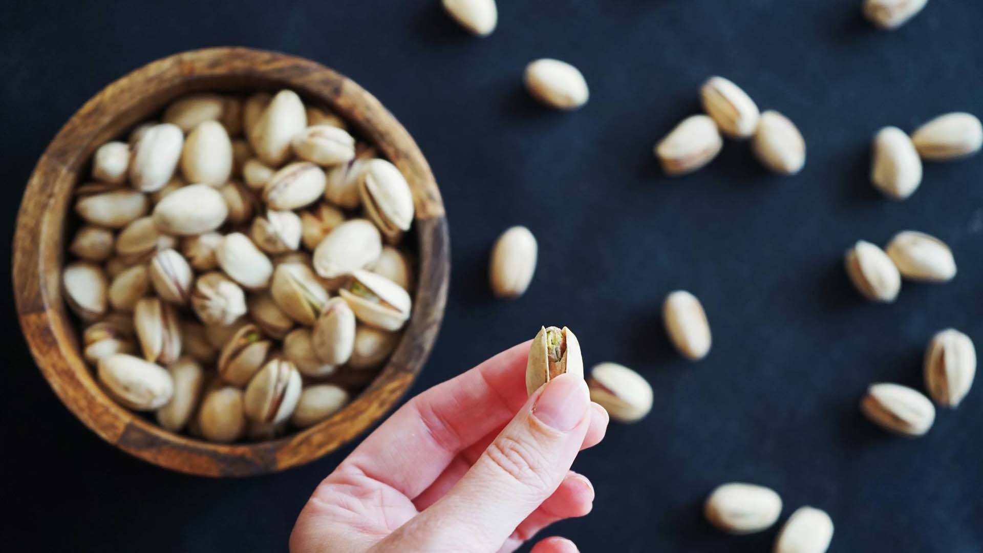 a close up of a hand holding a pisctachio nut with a bowl of them behind on a black background