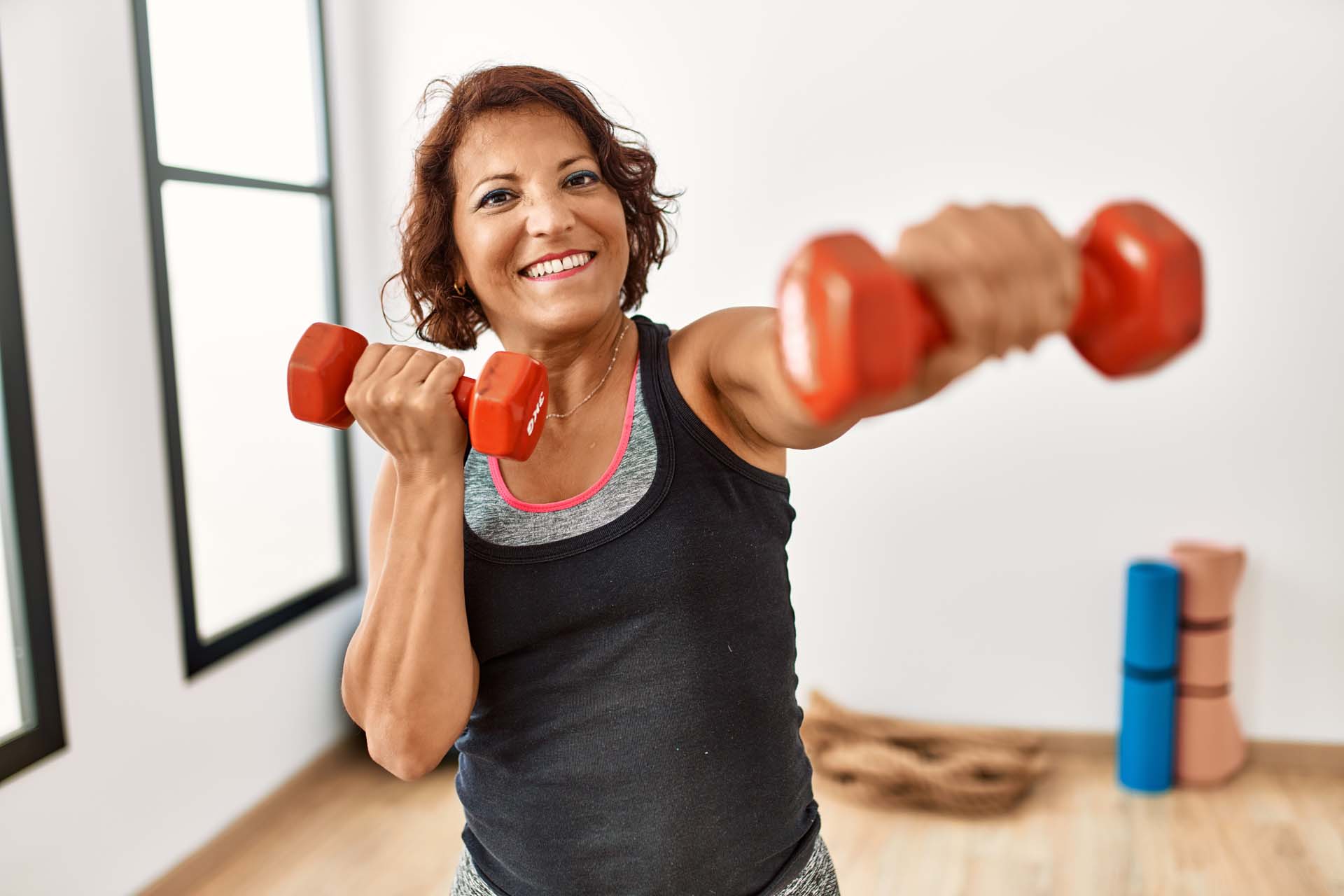 A smiling woman with a dumb bell in each hand in a gym