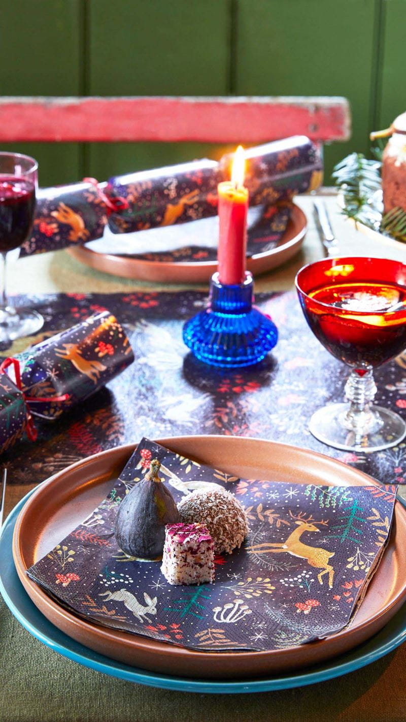 A festive table setting with matching napkins, cracker and table runner, a red candle in a blue candle holder and bright wine glasses