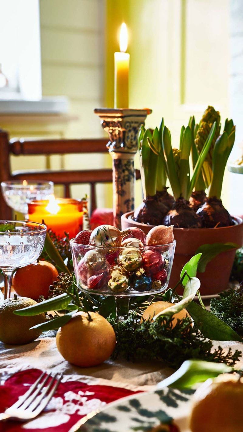 A festive table setting with a bowl of baubles in the centre piece, a tall candle stick with a lit candle and various fruits decorating the table