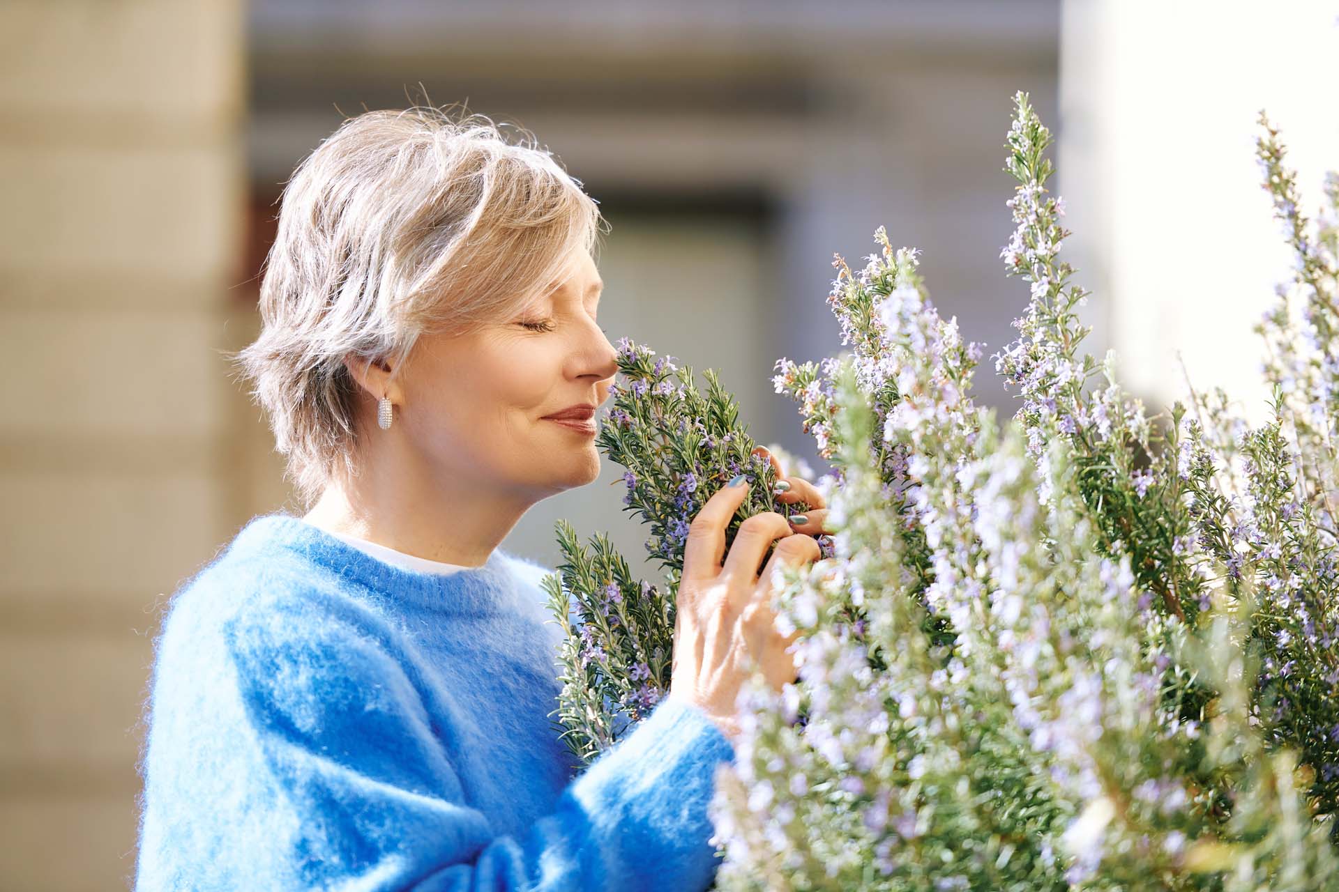 A woman in a blue jumper sniffing the flowers of a rosemary bush outside her home