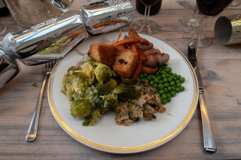 A plated Christmas dinner complete with turkey, pigs in blankets, roast potatoes, peas and Brussels sprouts, with a silver cracker next to the plate