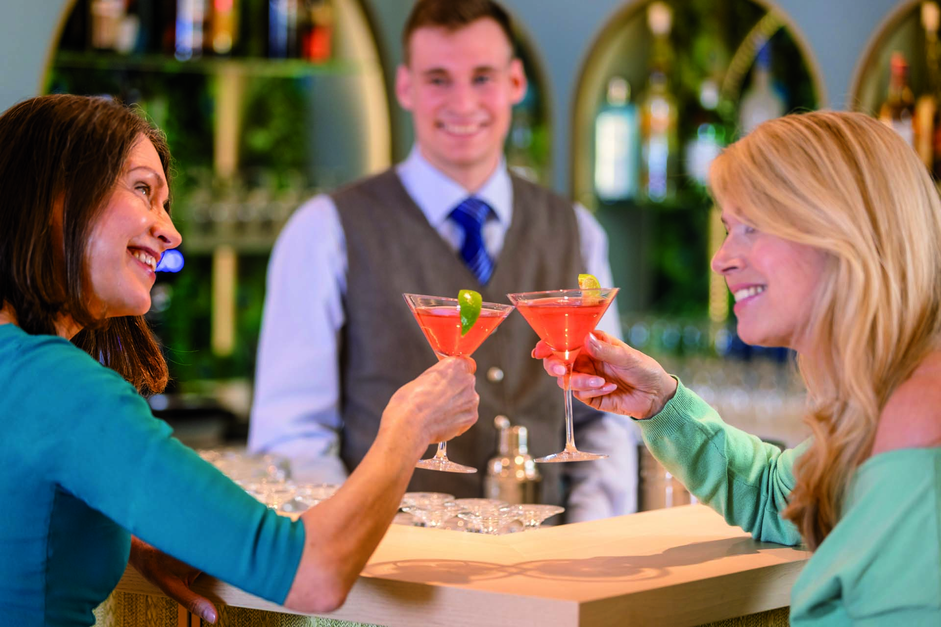 Two women at a bar with the bartender watching chinking their orange cocktails and looking happy