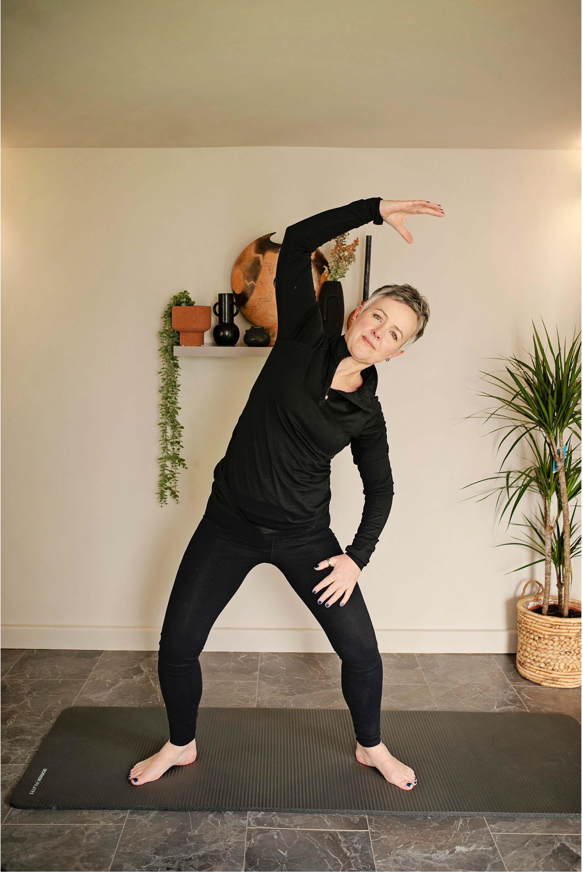 A woman demonstrating a standing mermaid stretch with one arm arching over her head
