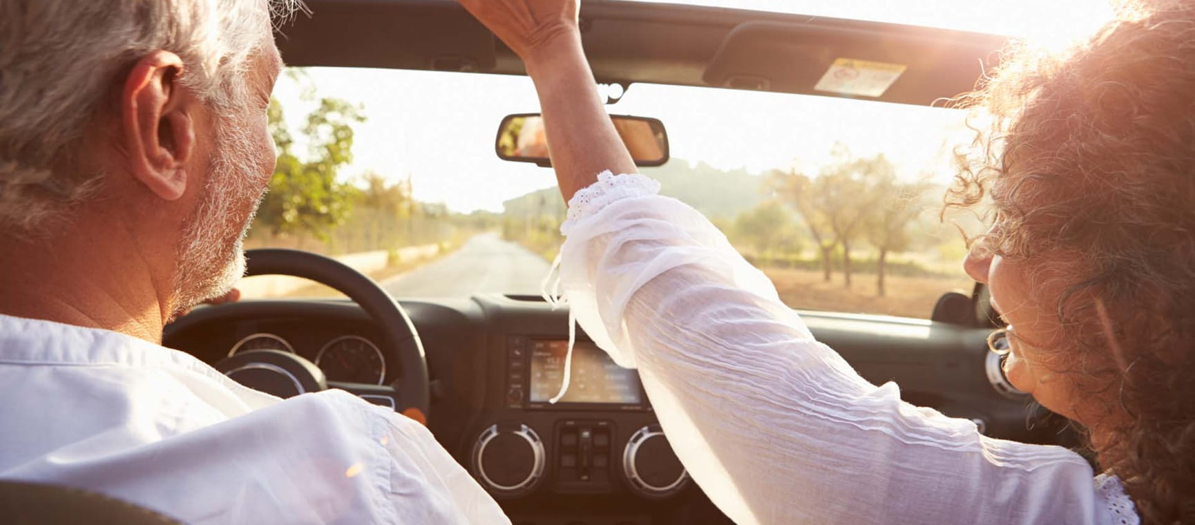 An older couple driving a convertible through sunny landscape, with the woman with her hands in the air and the man smiling