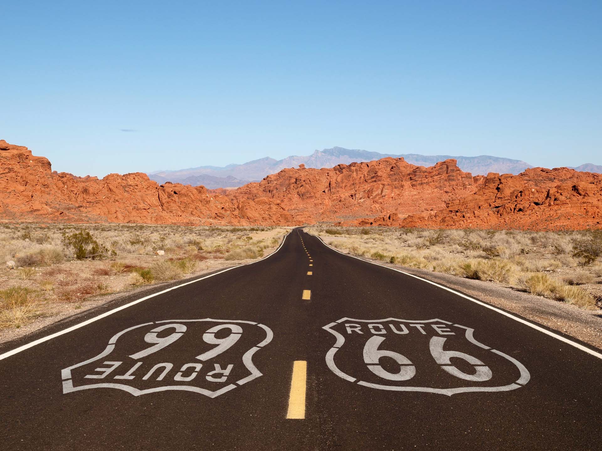 A long road stretching off into the distance with far off dessert mountains, and the iconic symbol of Route 66 on the tarmac