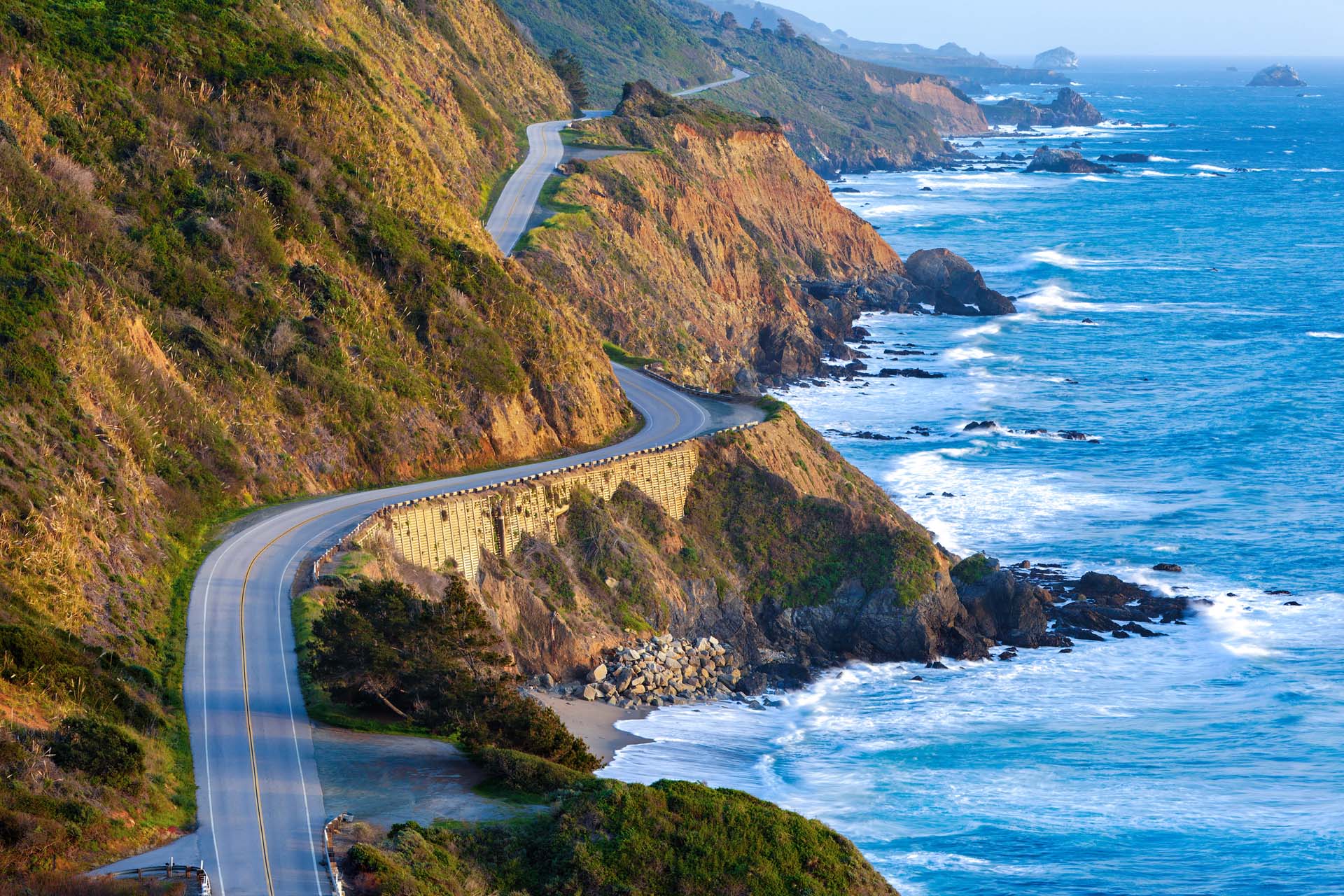 A road winding along the Pacific coast of the USA, with crashing waves to the side.