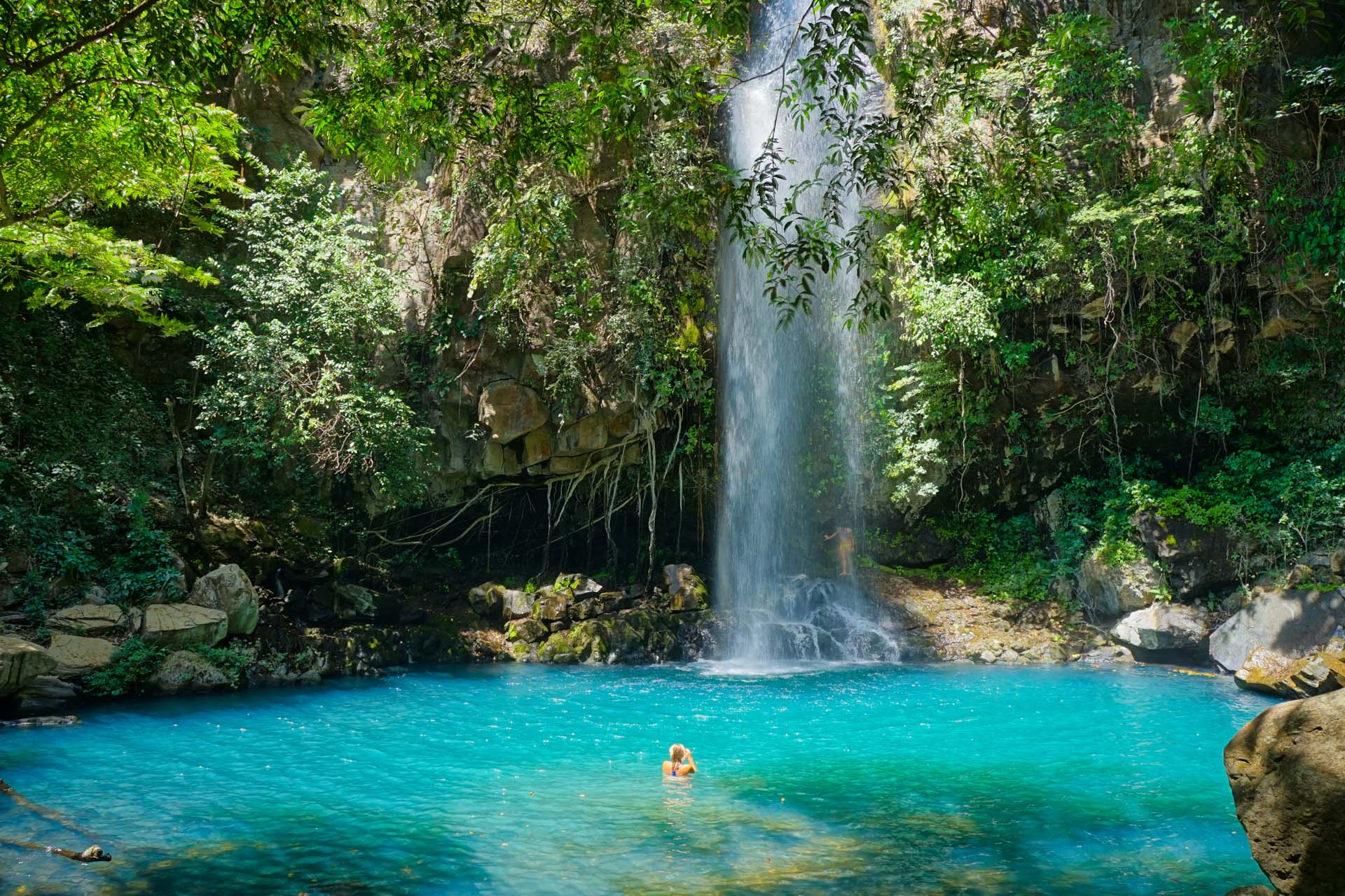 An idyllic waterfall in Costa Rica, with bright blue water and a swimmer. Surrounded by rocks and trees