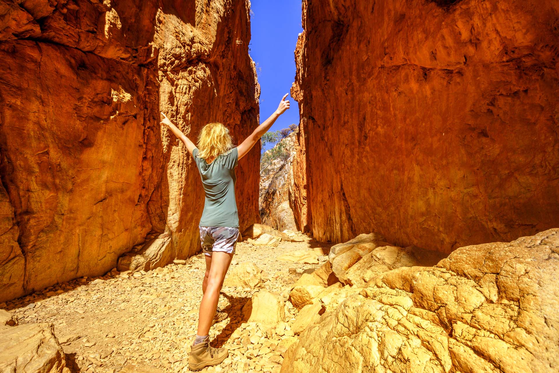 The back of a woman with long blonde hair standing with her arms raised in the middle of a gap between rock cliffs in the Australian desert