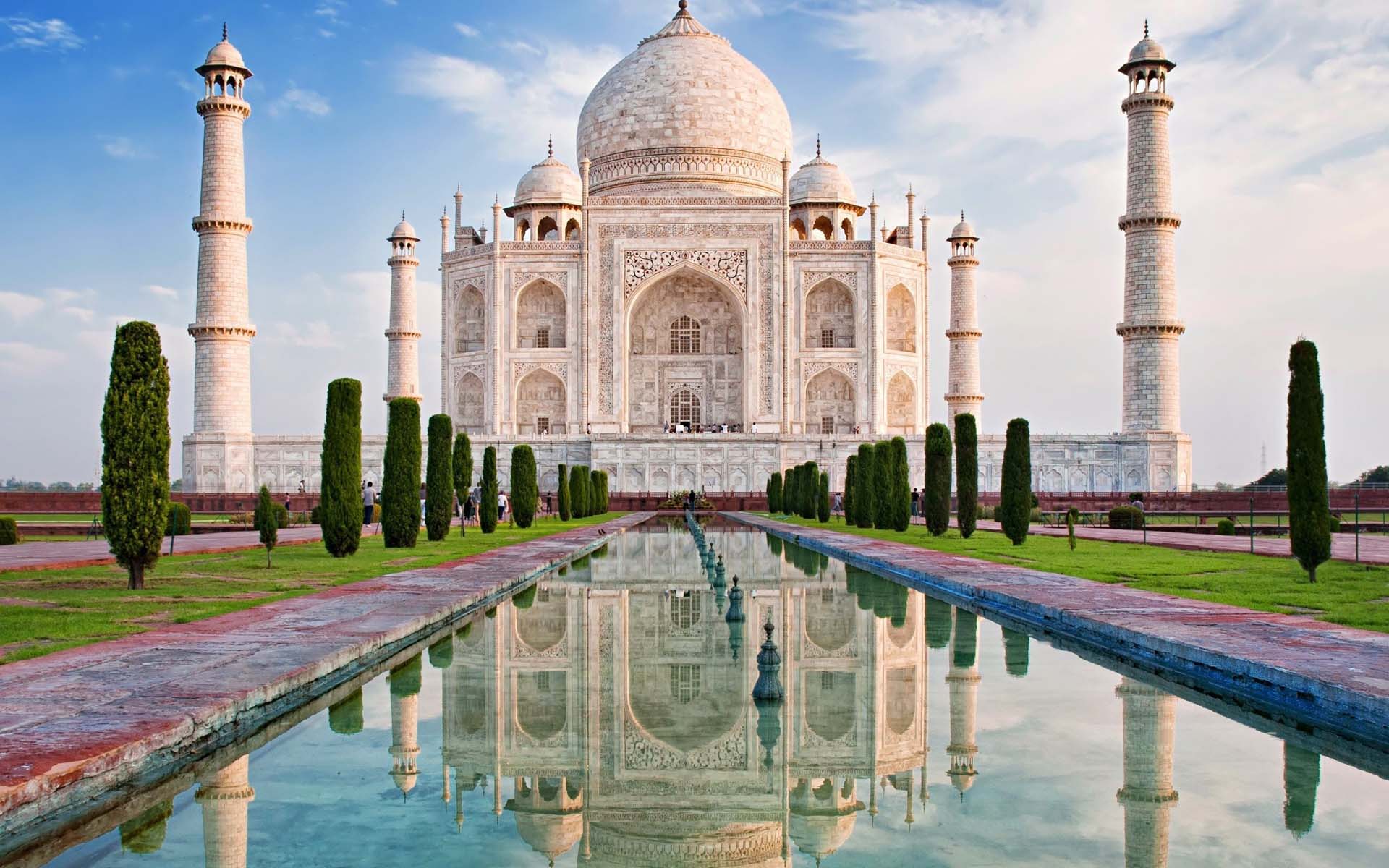 The ivory white marble front of the Taj Mahal building, in India, with its water feature in the foreground and blue sky behind