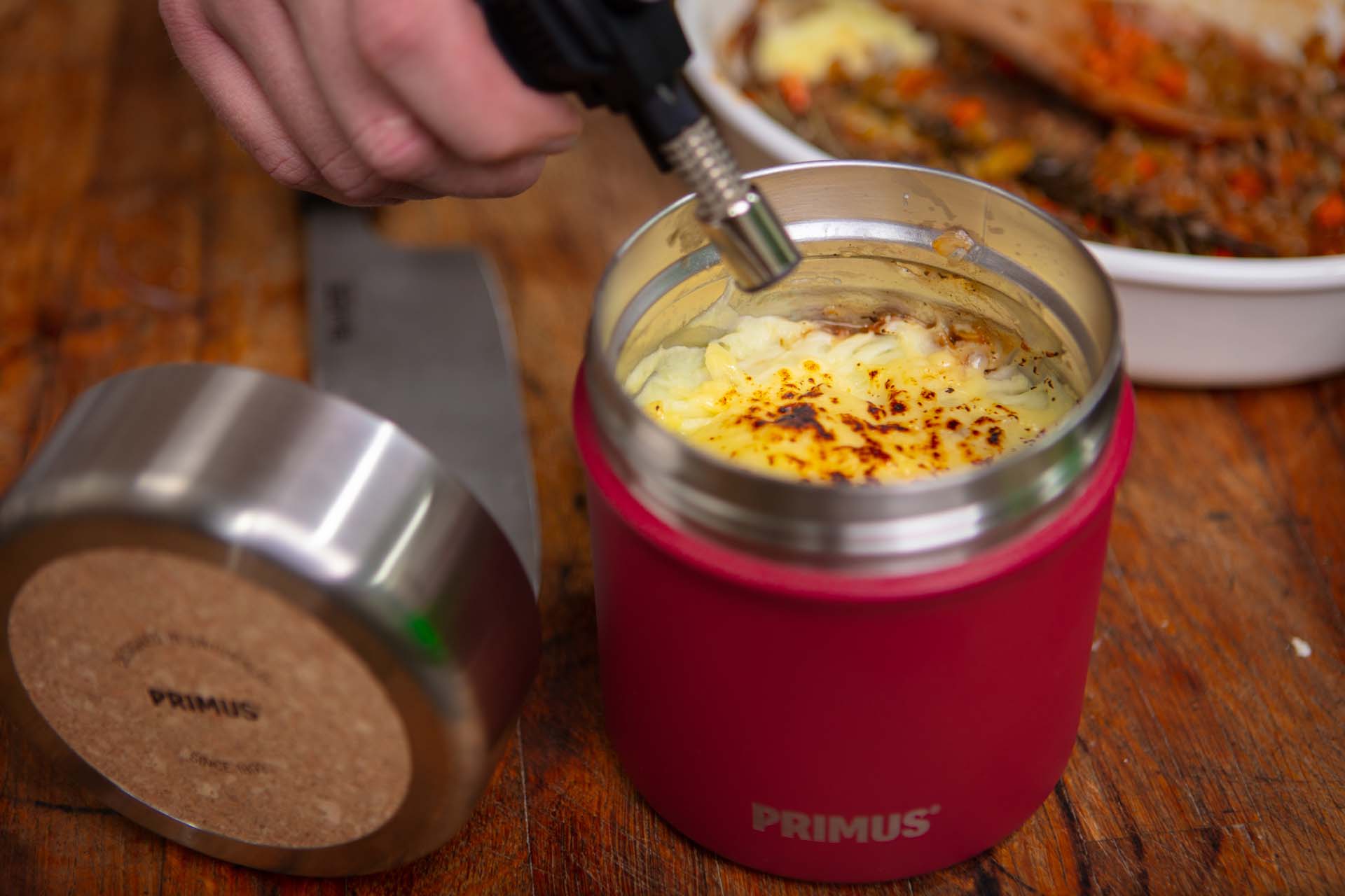 Close-up of someone using a blowtorch to brown the top of a shepherd's pie in a red food flask