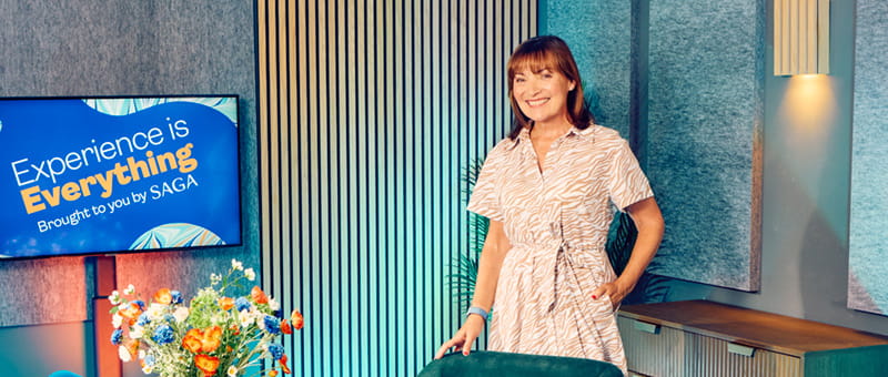 Person standing in a presentation space beside a screen displaying ‘Experience is Everything brought to you by Saga’, with flowers in the foreground.
