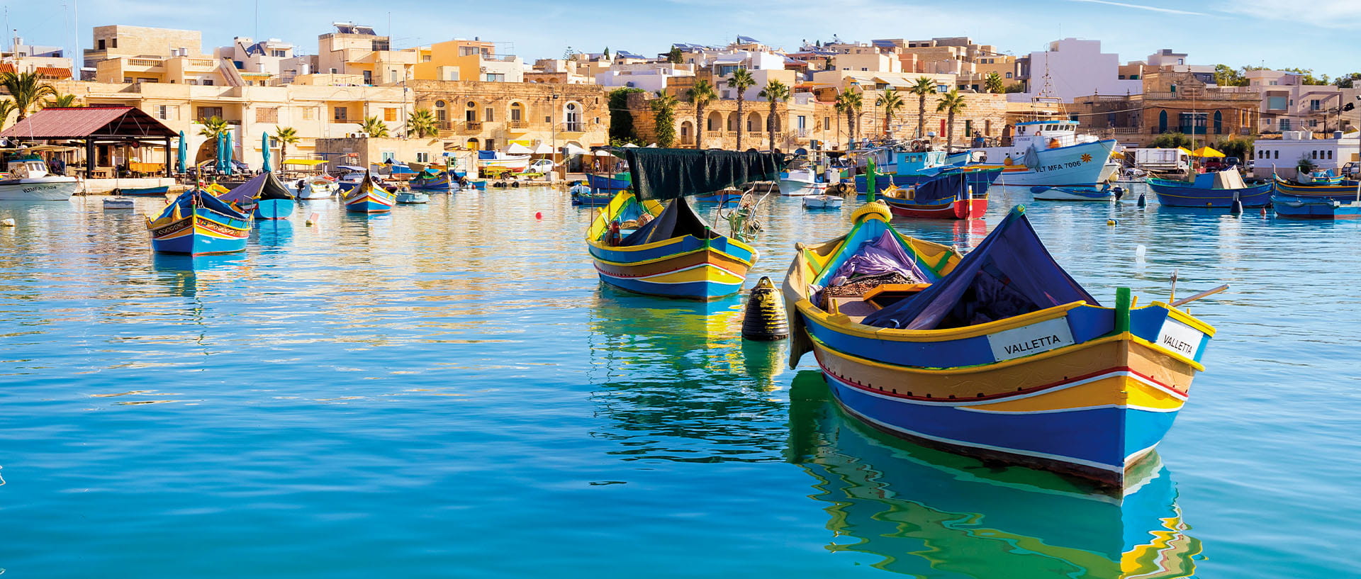 Colorful traditional fishing boats floating in a calm harbor, with a waterfront town and low-rise buildings in the background.