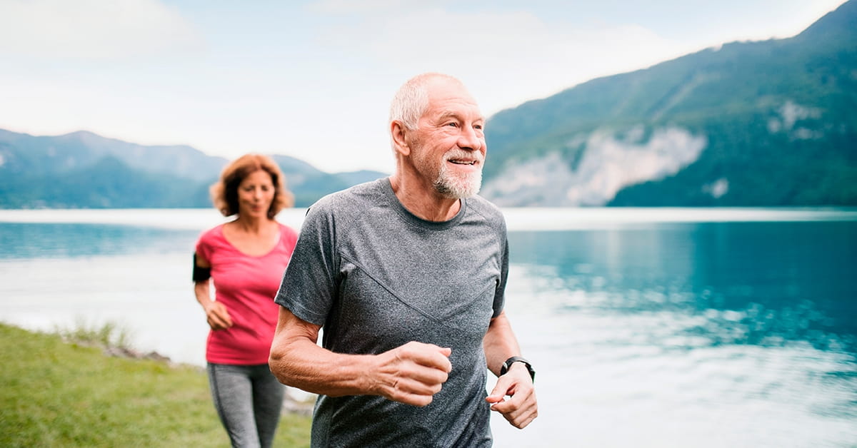 Two people jogging along a lakeside path with mountains and calm water in the background.