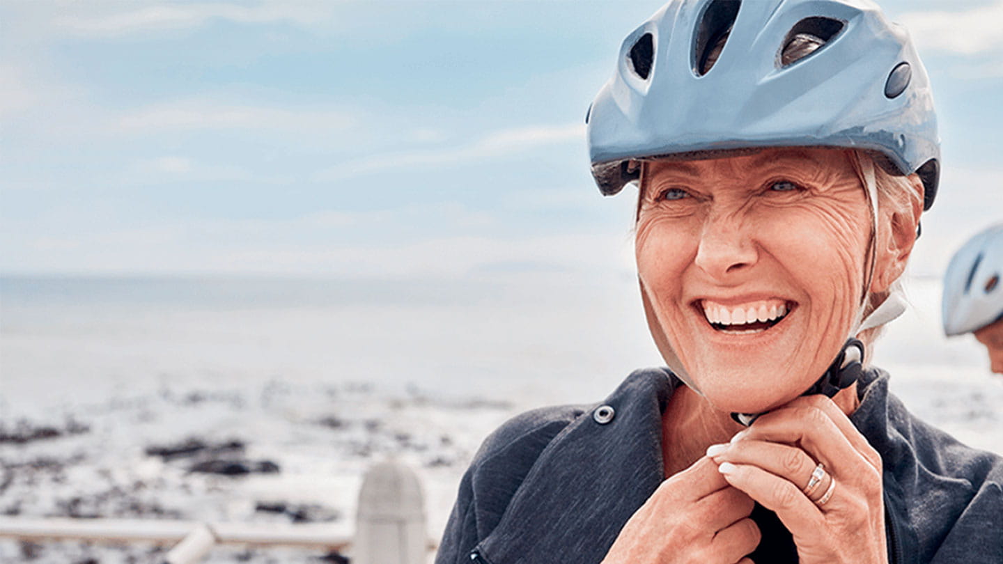 Person fastening a bicycle helmet near a coastal area with another cyclist in the background.