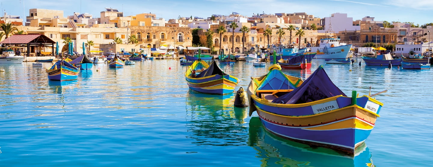 Colourful boats floating in a sunny harbour with a town in the background.
