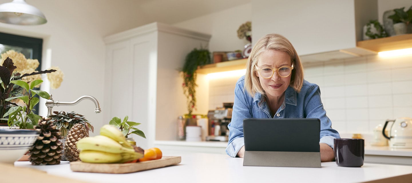 Mature woman at home in kitchen drinking coffee and looking at digital tablet