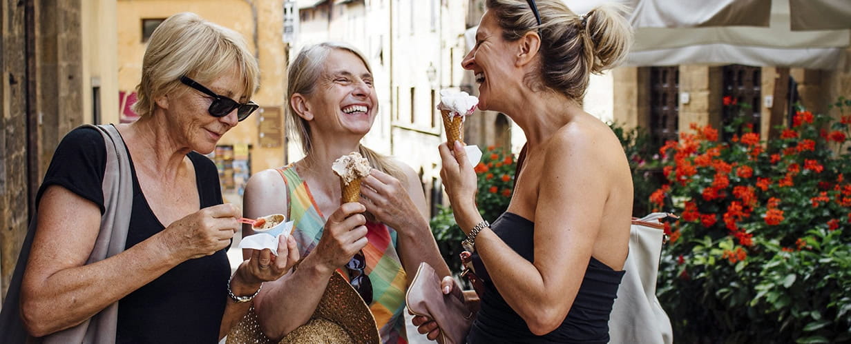 Three mature female friends standing eating Italian ice-creams while in a street in Tuscany during summer.