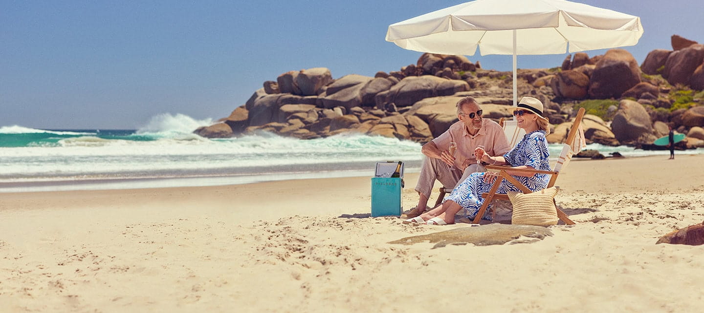 A mature couple sitting together under a parasol on a sandy beach.