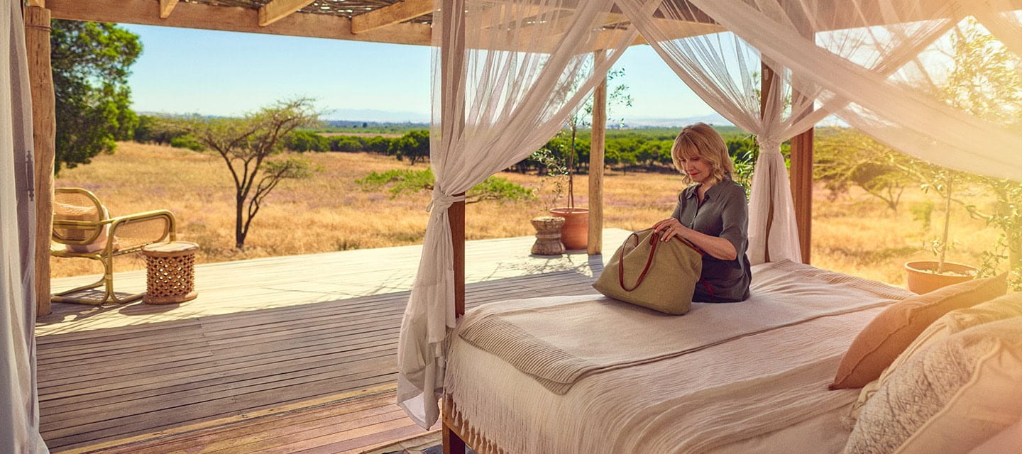 A mature woman is looking through her bag whilst sat on the bed of her safari resort hotel room.