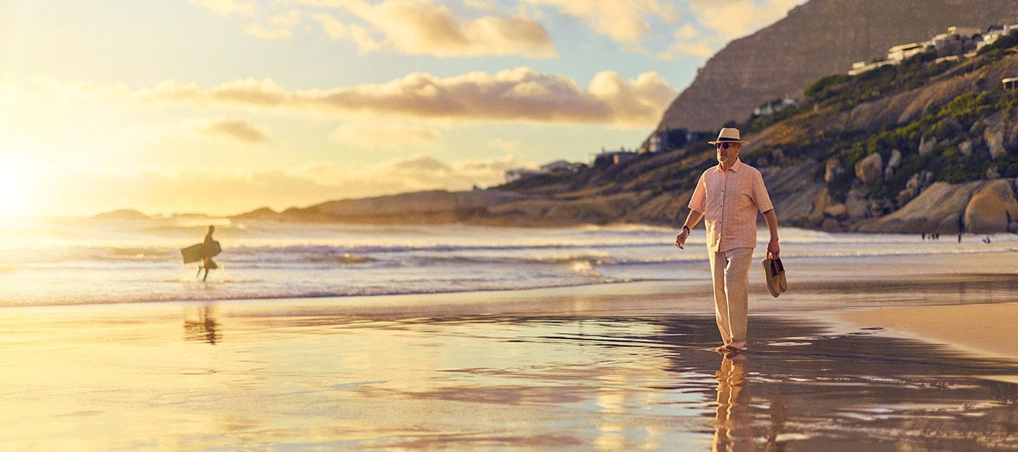 A mature man walking along a sandy beach at sunset holding his shoes.