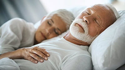 Closeup of mid 60's couple sleeping late in the morning in an already brightly lit bedroom.