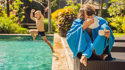 Poorly man caught a cold on holiday, sits sad at the pool drinking tea and blows his nose into a napkin. His grandson is healthy and swimming in the pool.