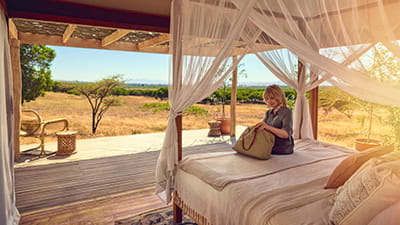 A mature woman is looking through her bag whilst sat on the bed of her safari resort hotel room.