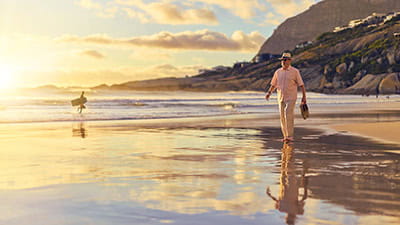 A mature man walking along a sandy beach at sunset holding his shoes.