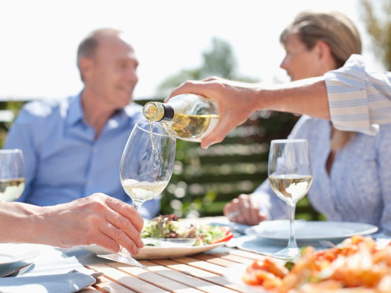 A glass of white wine being poured at an outdoor table with two adults chatting in the background and food on the table