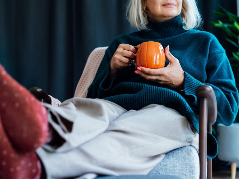 A woman wearing a navy knitted jumper and winter socks with her feet up, holding a pumpkin mug