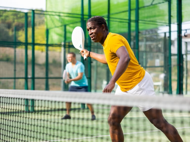A friendly doubles match of tennis; in focus is a man wearing a yellow t-shirt with tennis racket raised to receive the ball