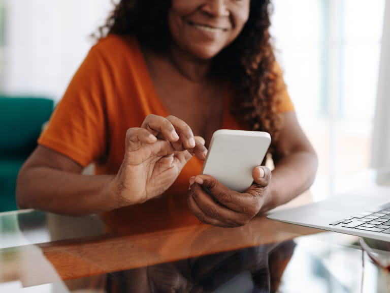 A woman with dark curly hair using a smartphone at a table, with a laptop visible to the side