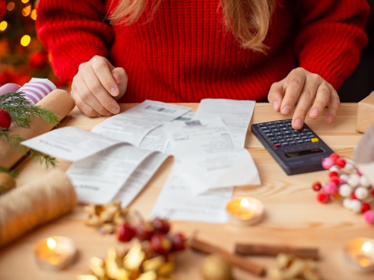 A woman in a red knitted jumper sitting at a desk which is covered in receipts, a calculator and Christmas decorations