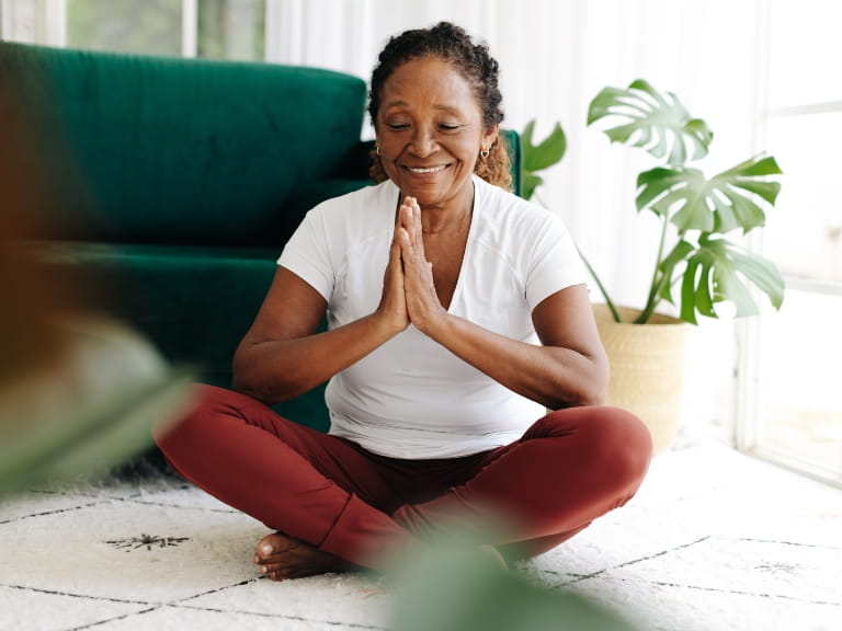 A woman sitting cross-legged with her palms together at her chest, practising yoga in a living room surrounded by house plants