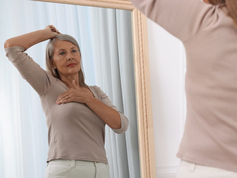 An older woman performing a health check under her arm in front of a mirror