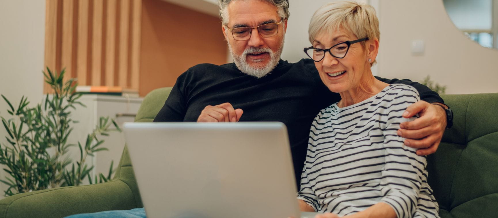 An older man and woman sat on a sofa looking at laptop together and smiling