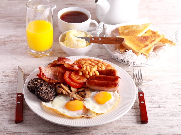 A full English breakfast with fried eggs, sausages, black pudding, beans, bacon, tomatoes and mushrooms, with a glass of orange juice and buttered toast on the side